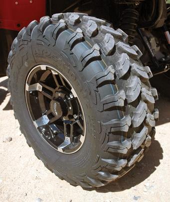 Close-up of a rugged off-road tire mounted on a black wheel, showing detailed tread pattern, parked on a dusty surface.