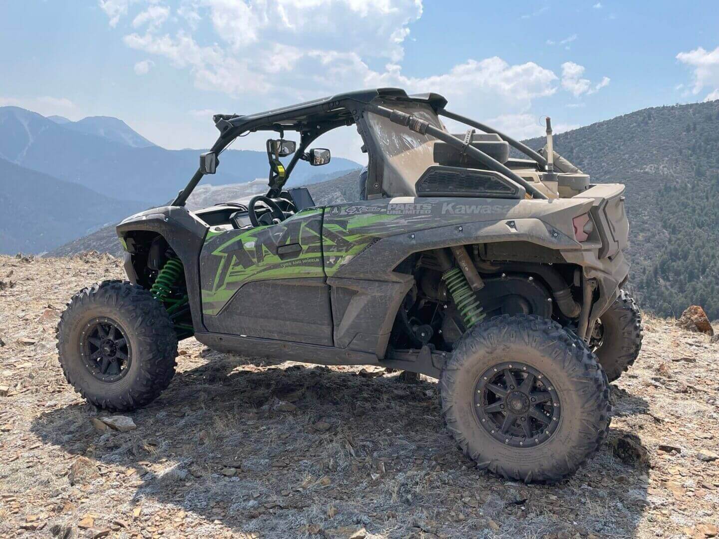 A side-profile image of a dusty kawasaki off-road vehicle parked on a rocky hillside with mountainous terrain in the background.