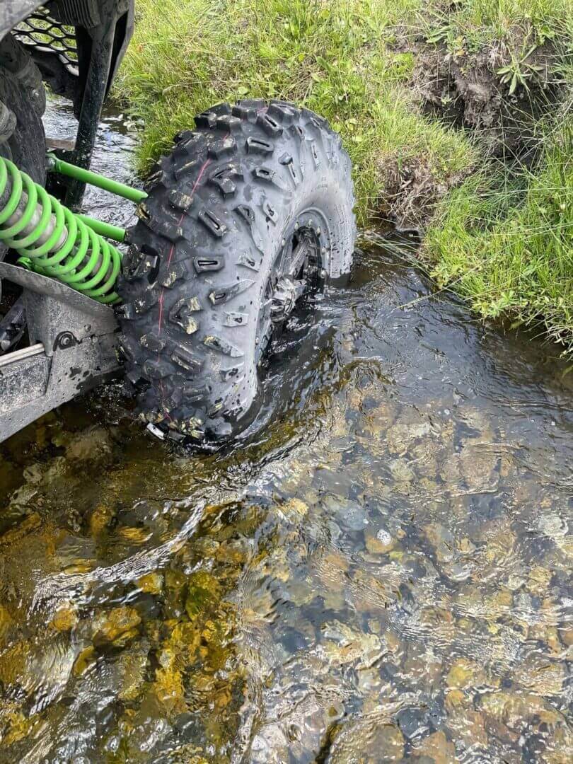 Atv tire submerged in a clear stream, showing detail of tread and suspension springs, with grassy banks on the sides.