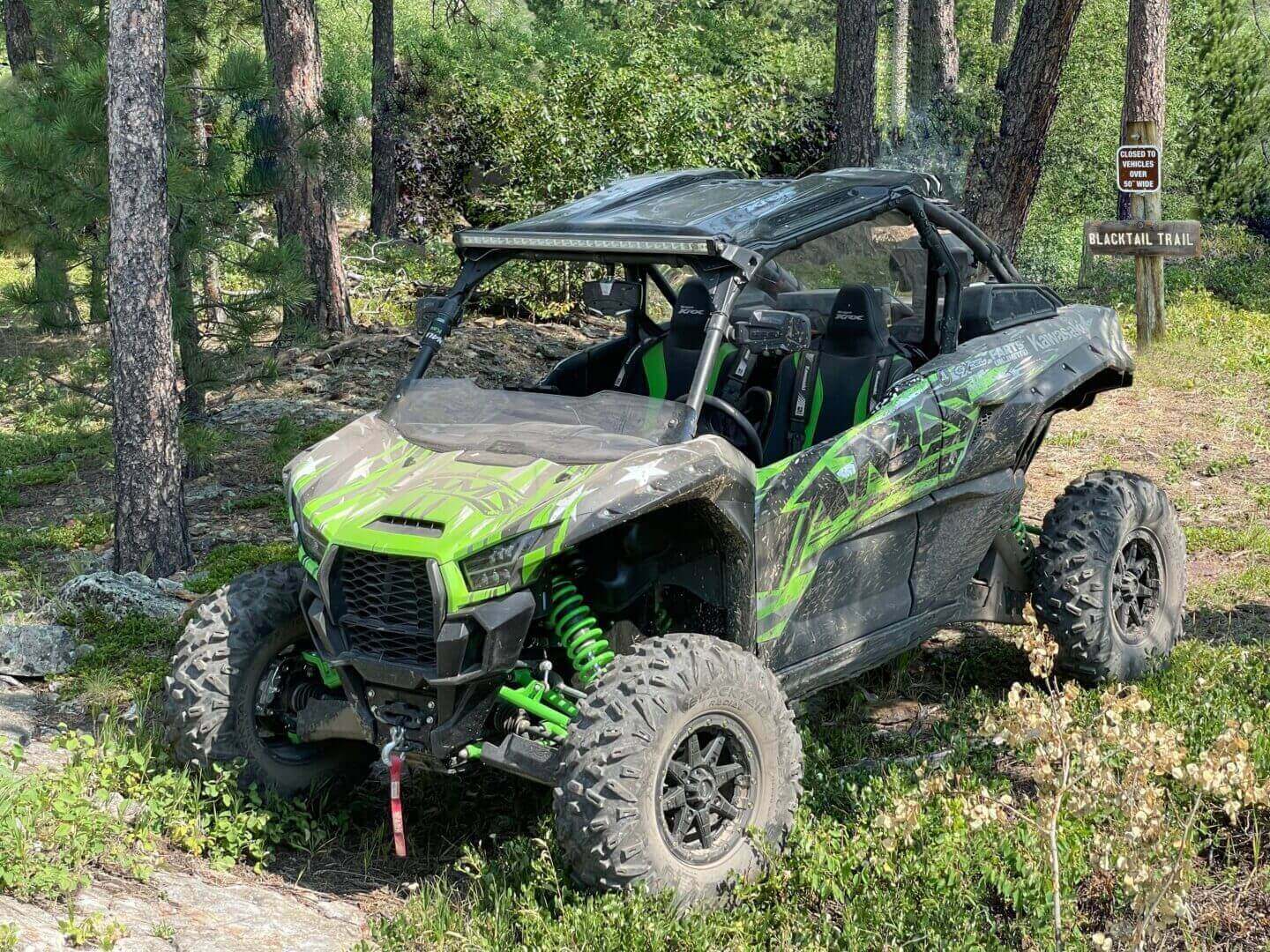 A green and black all-terrain vehicle parked on a forest trail, surrounded by pine trees.