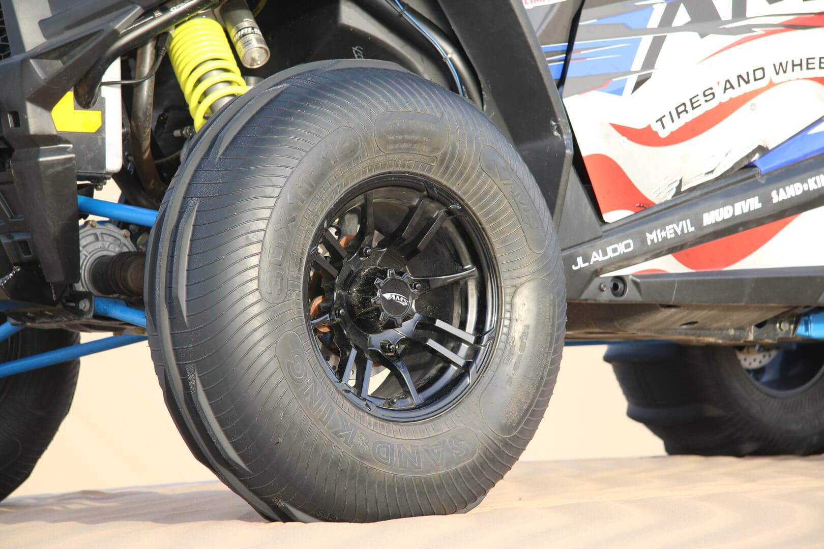 Close-up of a black, sandy off-road vehicle tire with visible suspension components and part of the vehicle's bodywork, taken during the daytime.