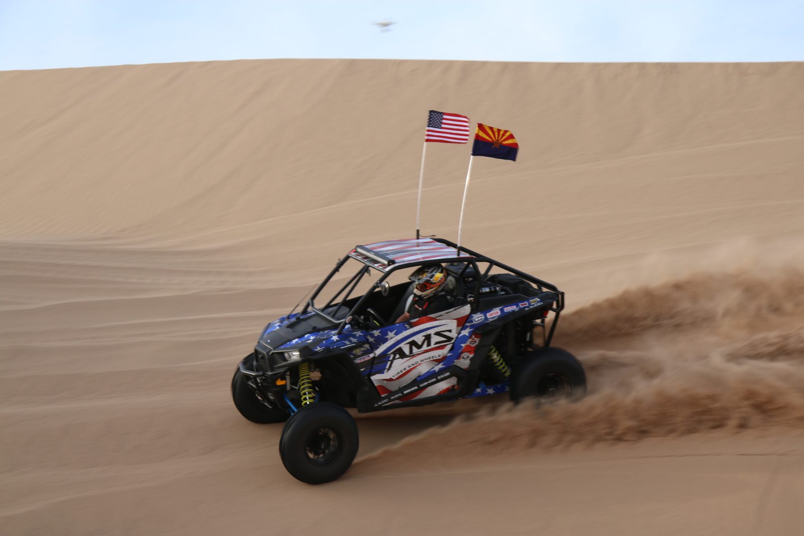 A blue off-road vehicle speeds across a sandy dune, kicking up dust, with american and arizona state flags flying from the back.