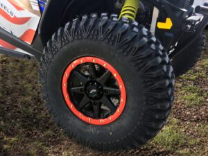 Close-up of an off-road vehicle tire with a red rim and black spokes, parked on grass, showing detailed tread pattern.