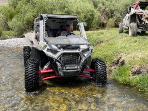 Man driving a four-wheel utility vehicle through a shallow river with grass and trees in the background.