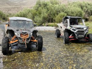 Two off-road vehicles parked in a shallow stream surrounded by grassy hills.