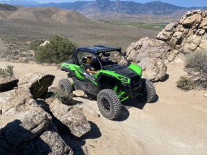 Two people in a green and black off-road vehicle navigating a rocky desert trail with mountains in the background.
