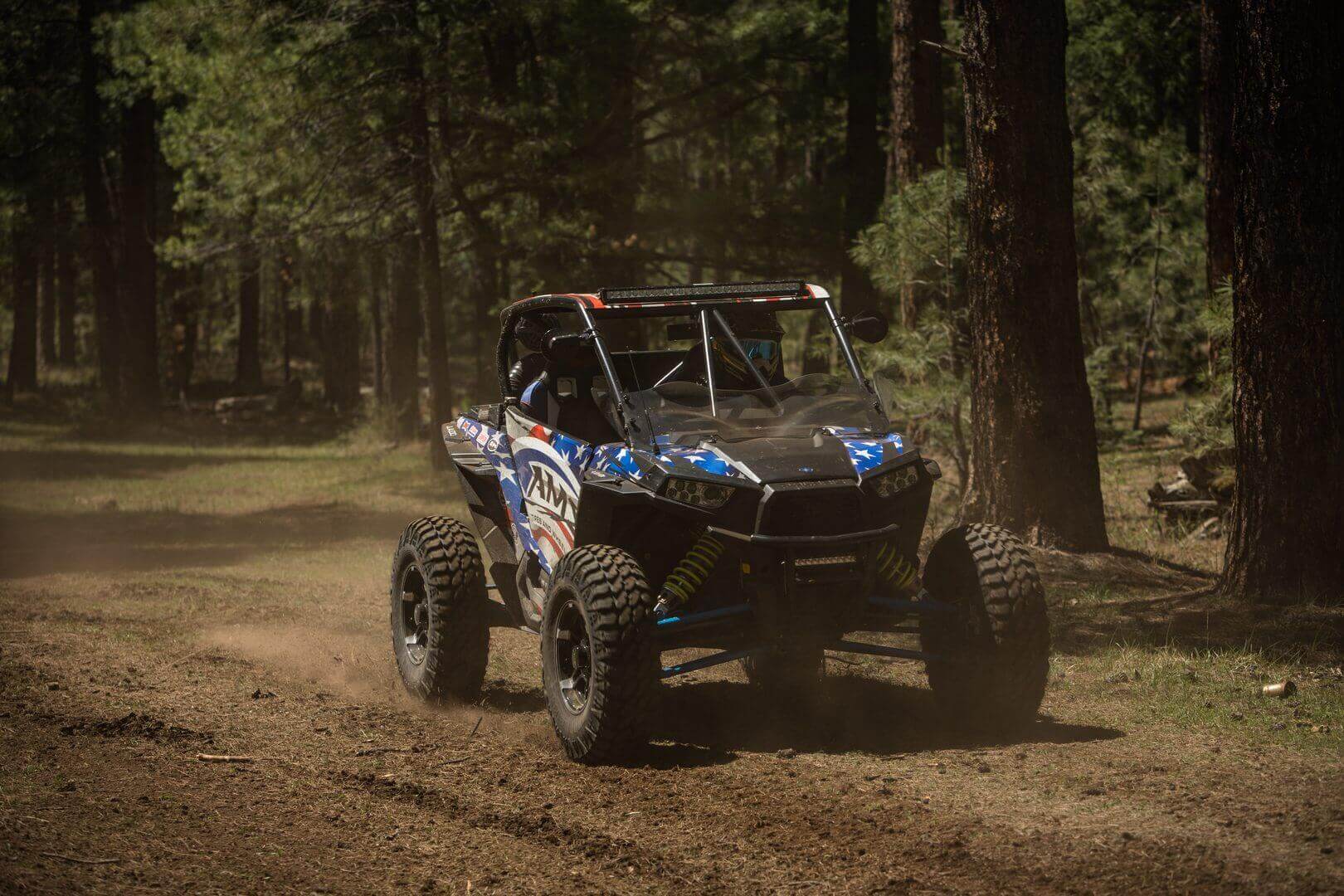 A blue and white off-road vehicle races through a dusty forest trail, its tires kicking up dirt among tall pine trees.
