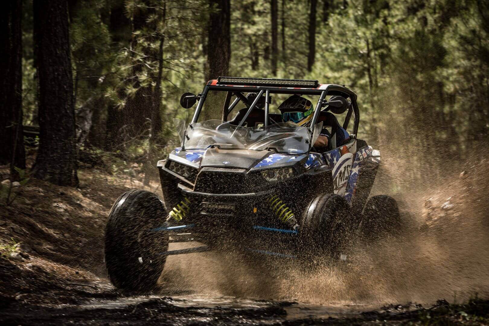 A person driving an off-road vehicle fast through a forested trail, kicking up dust and debris.