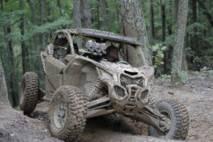 Person driving a muddy atv through a forest, navigating between trees on a rugged trail.